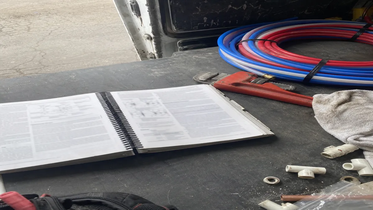 A spiral-bound plumbing code manual open on a van floor next to a pipe wrench, coiled PEX tubing, and scattered fittings