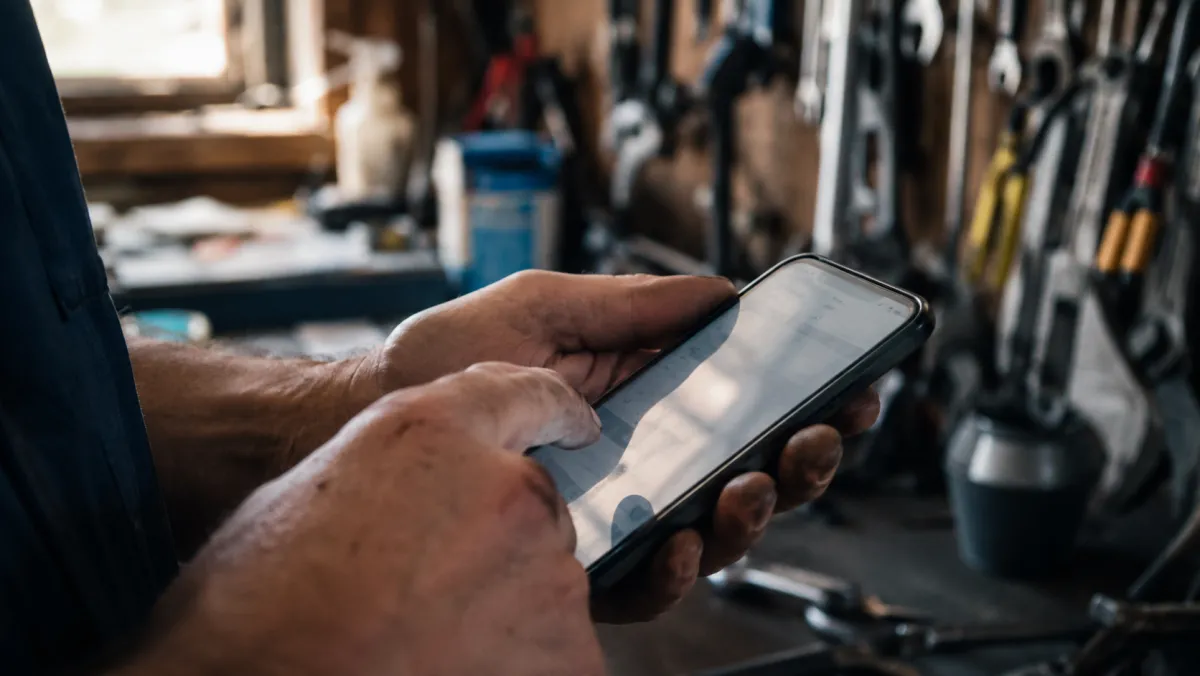 A plumber's hands tapping a smartphone in a tool-lined workshop