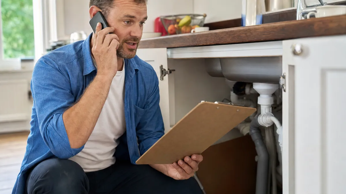 A plumber on the phone at a kitchen sink with a clipboard, talking a customer through pricing