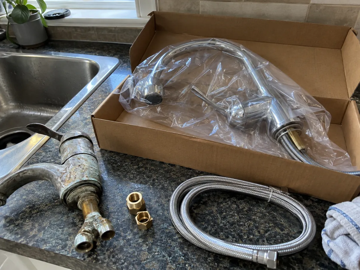 A new chrome kitchen faucet in an open cardboard box on a counter beside the corroded faucet it will replace