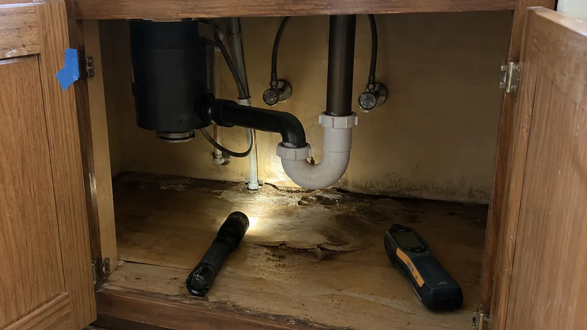 An open kitchen sink cabinet with water-stained flooring, a flashlight and a moisture meter, and a strip of blue tape on the door